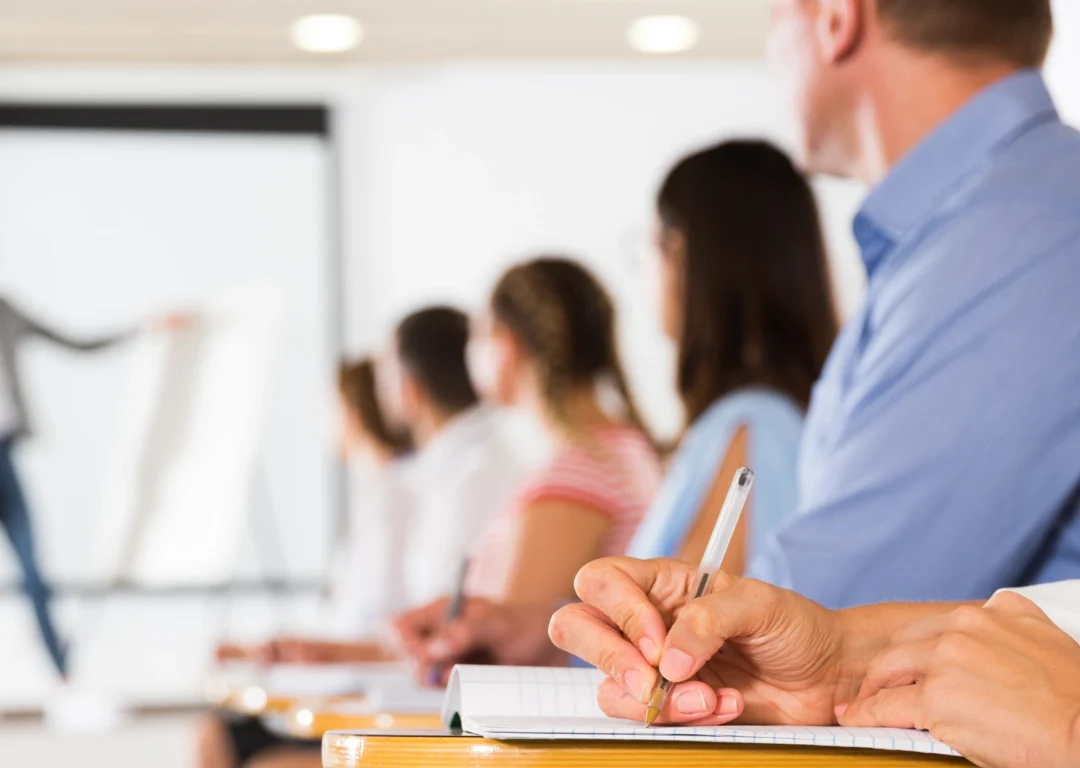 Men and women sat at individual wooden desks taking notes and listening to speaker next to whiteboard.