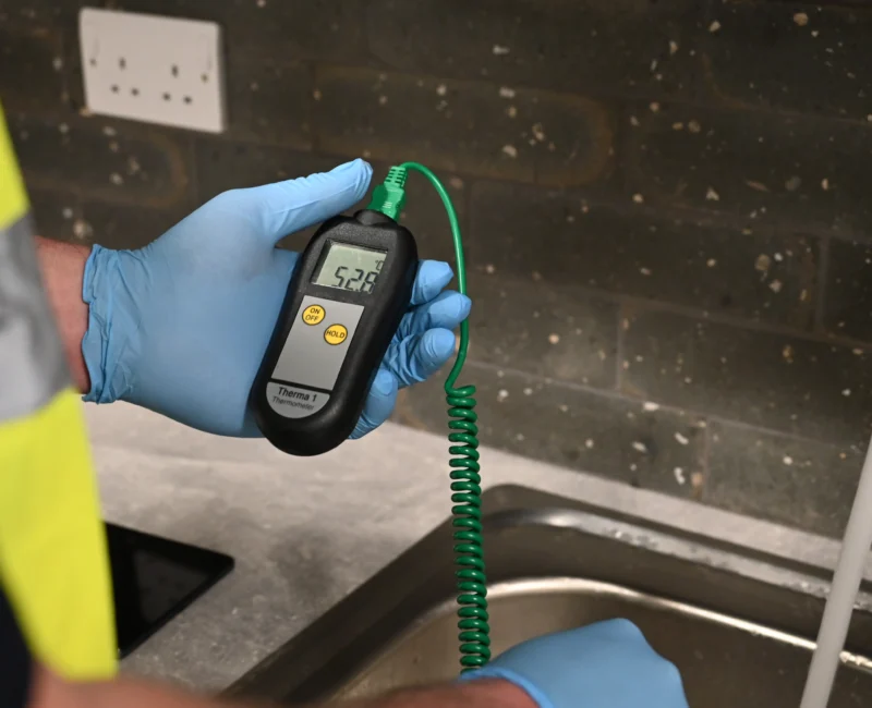 A person wearing blue protective gloves holds a digital thermometer with a green probe, measuring the temperature of water flowing from a tap.