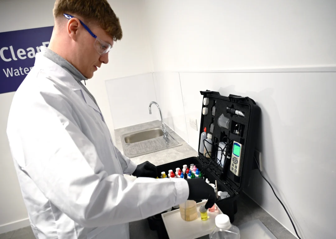 A male technician in a lab coat and safety glasses uses a water testing kit.